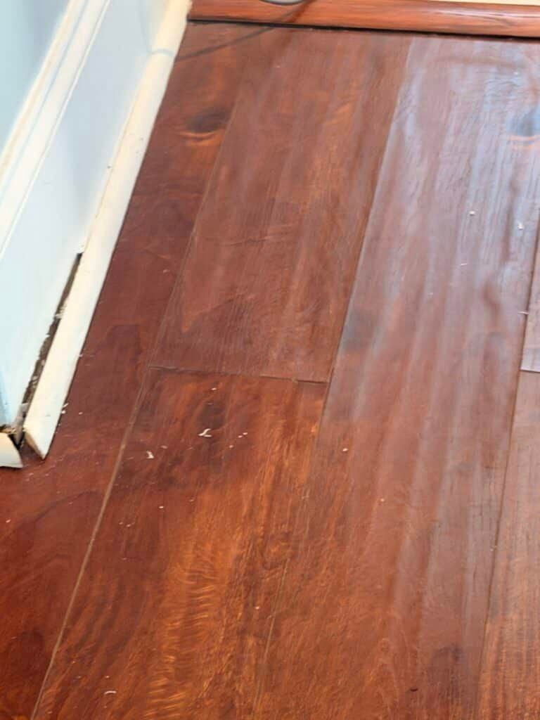 Discolored, graying hardwood floor plank from water damage next to a doorway threshold in a Charlotte NC home, the kind of small-looking spot homeowners weigh against filing a claim